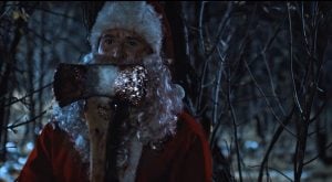 A person dressed as Santa Claus holds a blood-stained axe in a dark, wooded area at night.