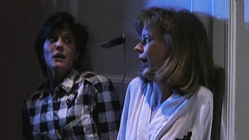 Two women sit against a wall looking frightened, with a knife protruding from the wall just above them.