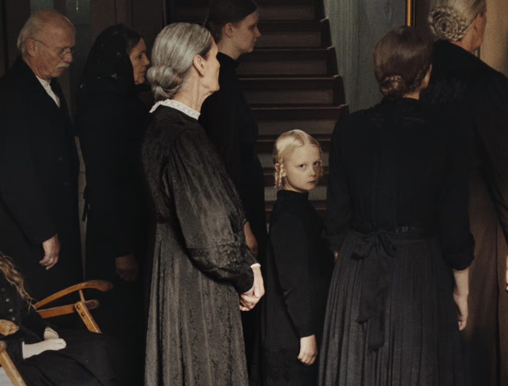 A group of people dressed in black period clothing stand indoors near a staircase; a young blonde girl faces the camera while others face away.