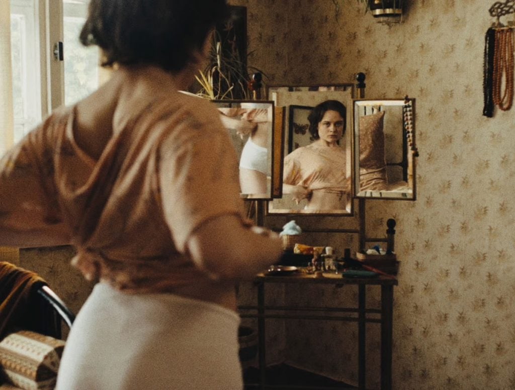 A woman with short dark hair stands in front of a vanity mirror, adjusting her shirt with her reflection visible. The room has patterned wallpaper and vintage decor.
