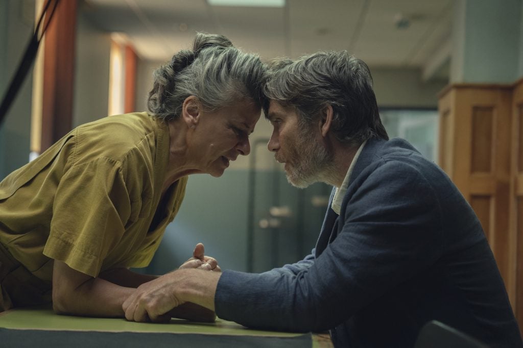 A woman in a prison uniform leans across a table, touching foreheads with a man in a suit during an emotional conversation in a visitation room.