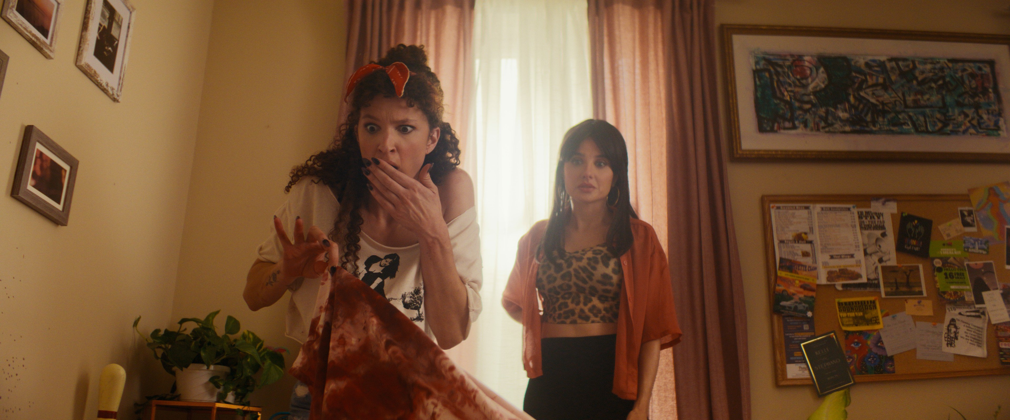 Two young women stand in a bedroom, one looking shocked with her hand over her mouth, holding a stained garment; the other looks on calmly.