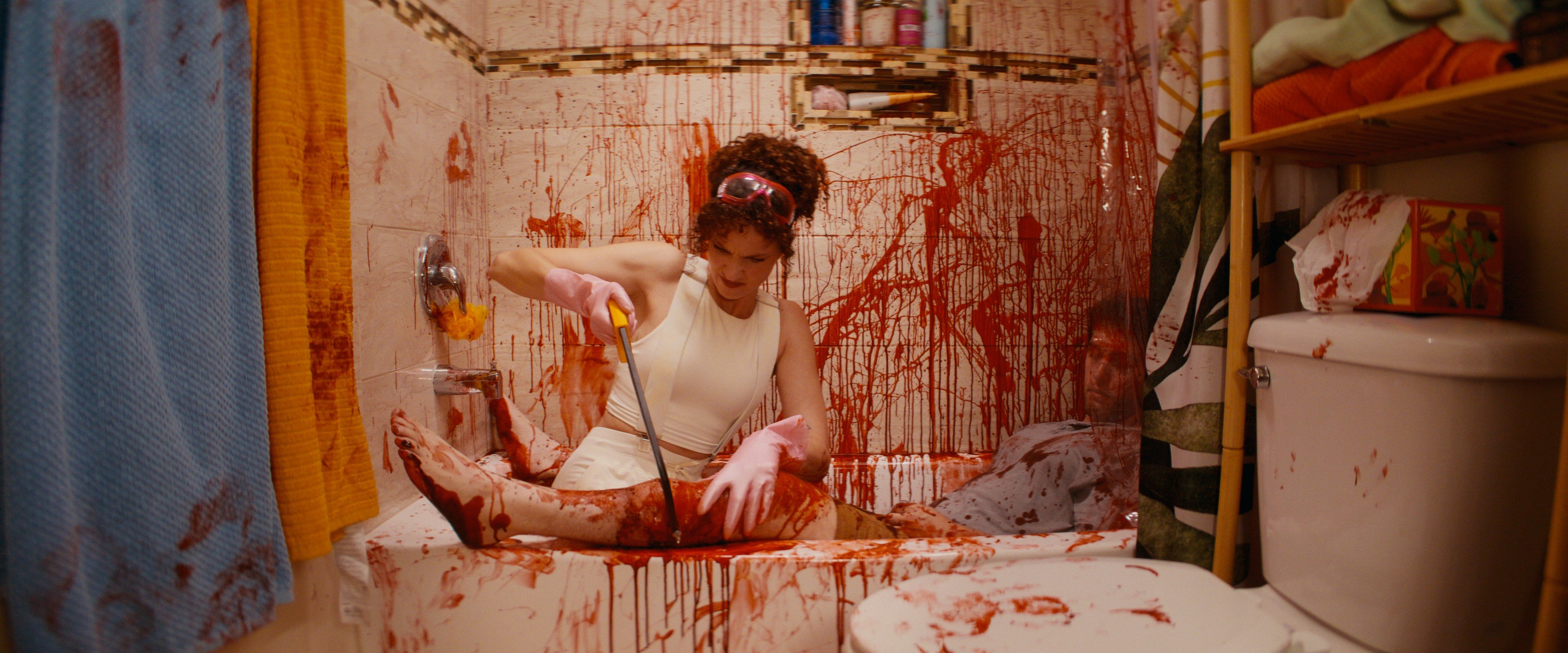 A woman in a white dress and pink gloves sits in a bathtub, using a saw on a body part amid a blood-splattered bathroom.