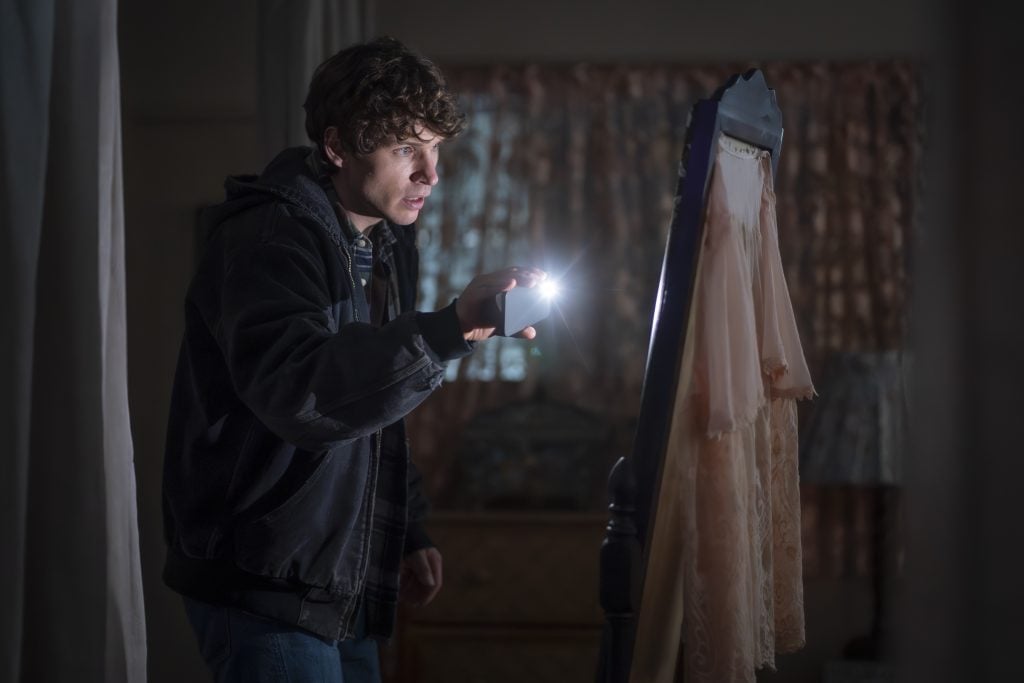 A young person in a dark room shines a flashlight onto a nightgown hanging on a standing mirror, looking at it with a focused expression.