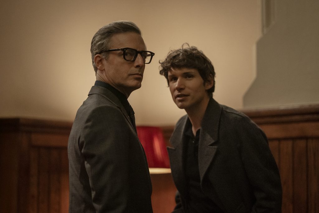 Two men in suits stand indoors near a wooden wall and a red lamp, both looking toward the camera with serious expressions.