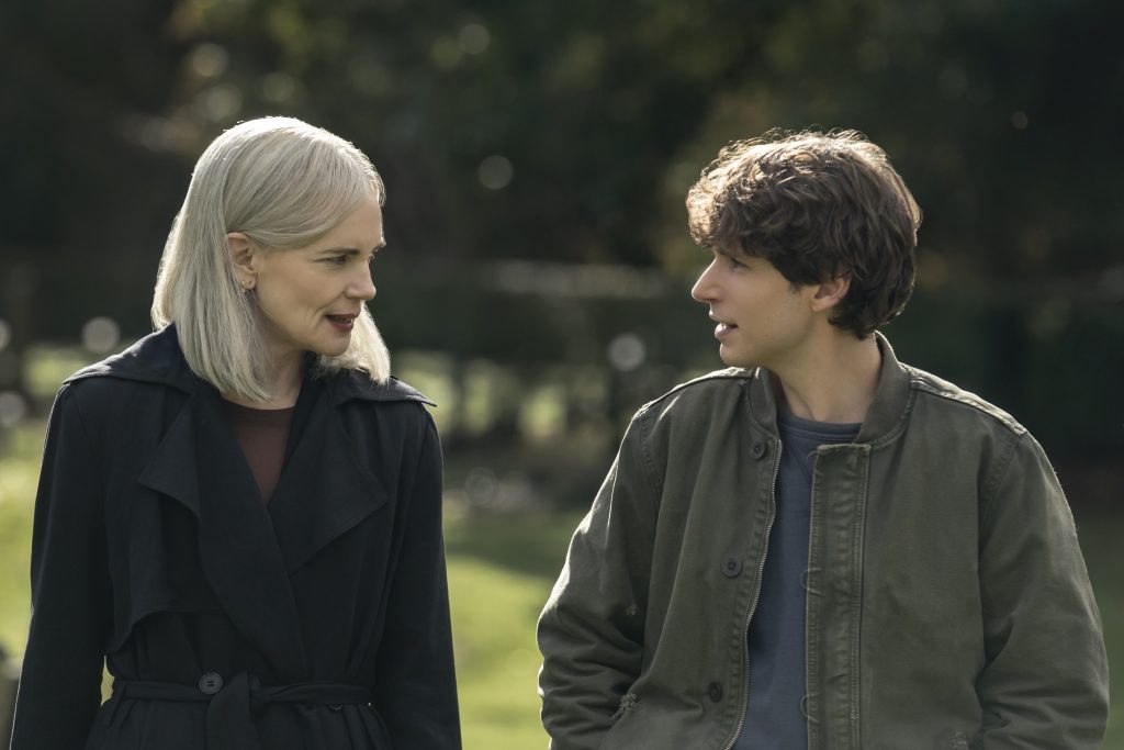 A woman with gray hair and a black coat walks outdoors beside a young man in a green jacket; both are engaged in conversation.