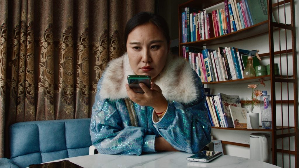 A woman in a blue sequined jacket uses her smartphone at a table, with a bookshelf and patterned curtains in the background.