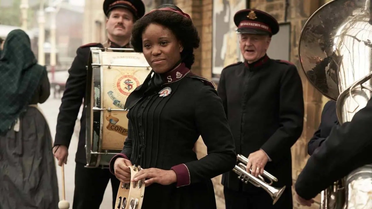 A woman in a uniform holds a tambourine, standing among other uniformed musicians with brass and percussion instruments on a street.