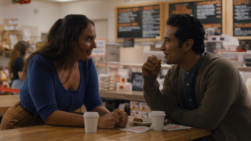 Two people sit at a café table with coffee and pastries, engaged in conversation. Menus and baked goods are visible in the background.