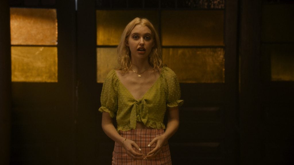 A woman with blonde hair wearing a green blouse and plaid skirt stands indoors in front of dark wooden doors with yellow-tinted glass panels.