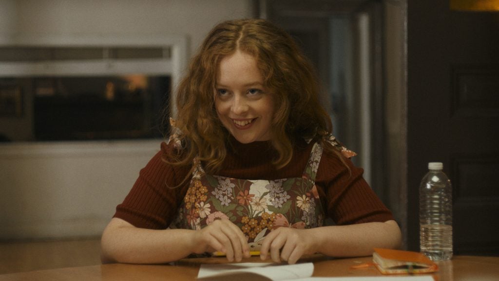 A young woman with curly red hair and a floral dress sits at a table, smiling and holding a pencil. A notebook and a bottle of water are on the table in front of her.