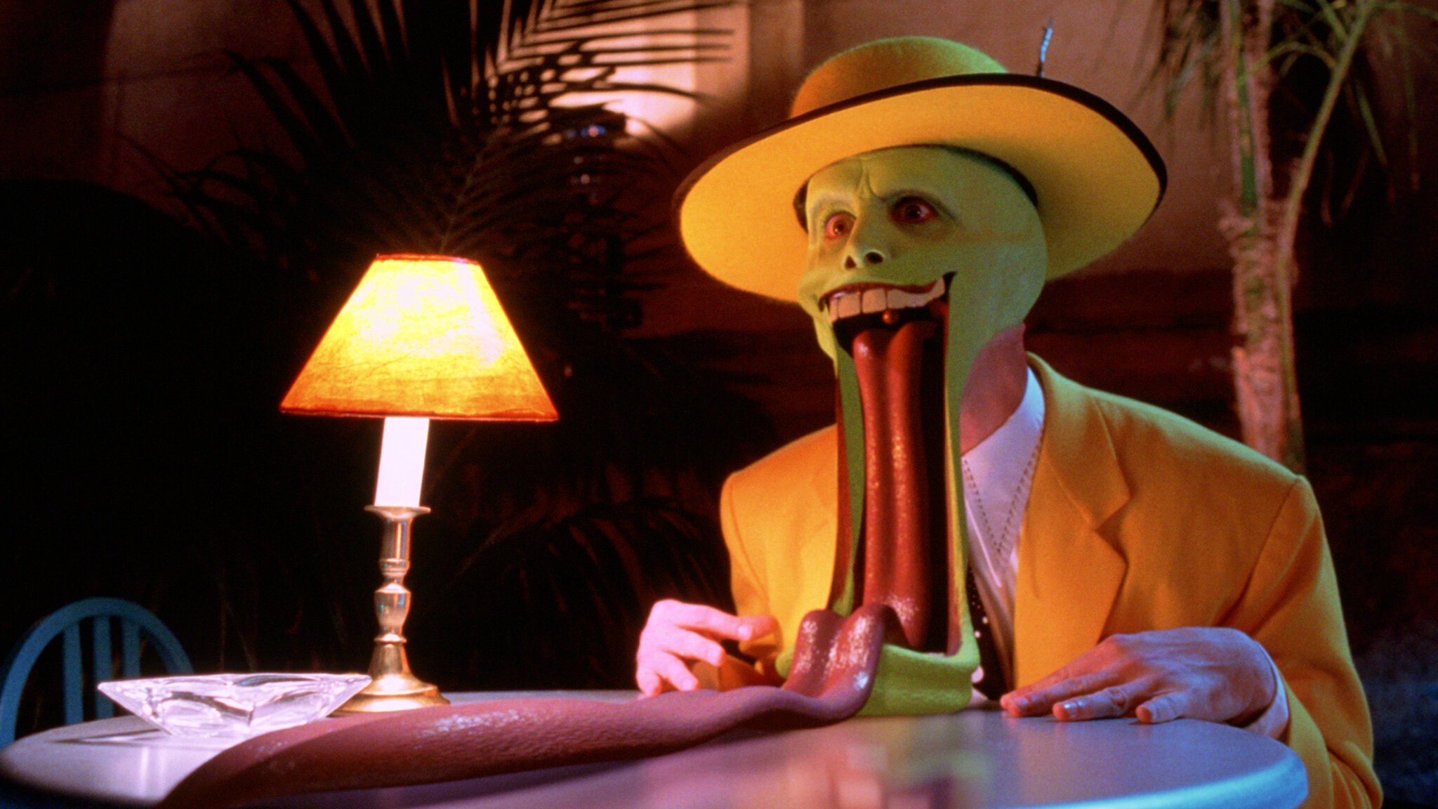 A person in a bright yellow suit and hat sits at a table, their green face stretched with a long tongue extended, under a small lamp.