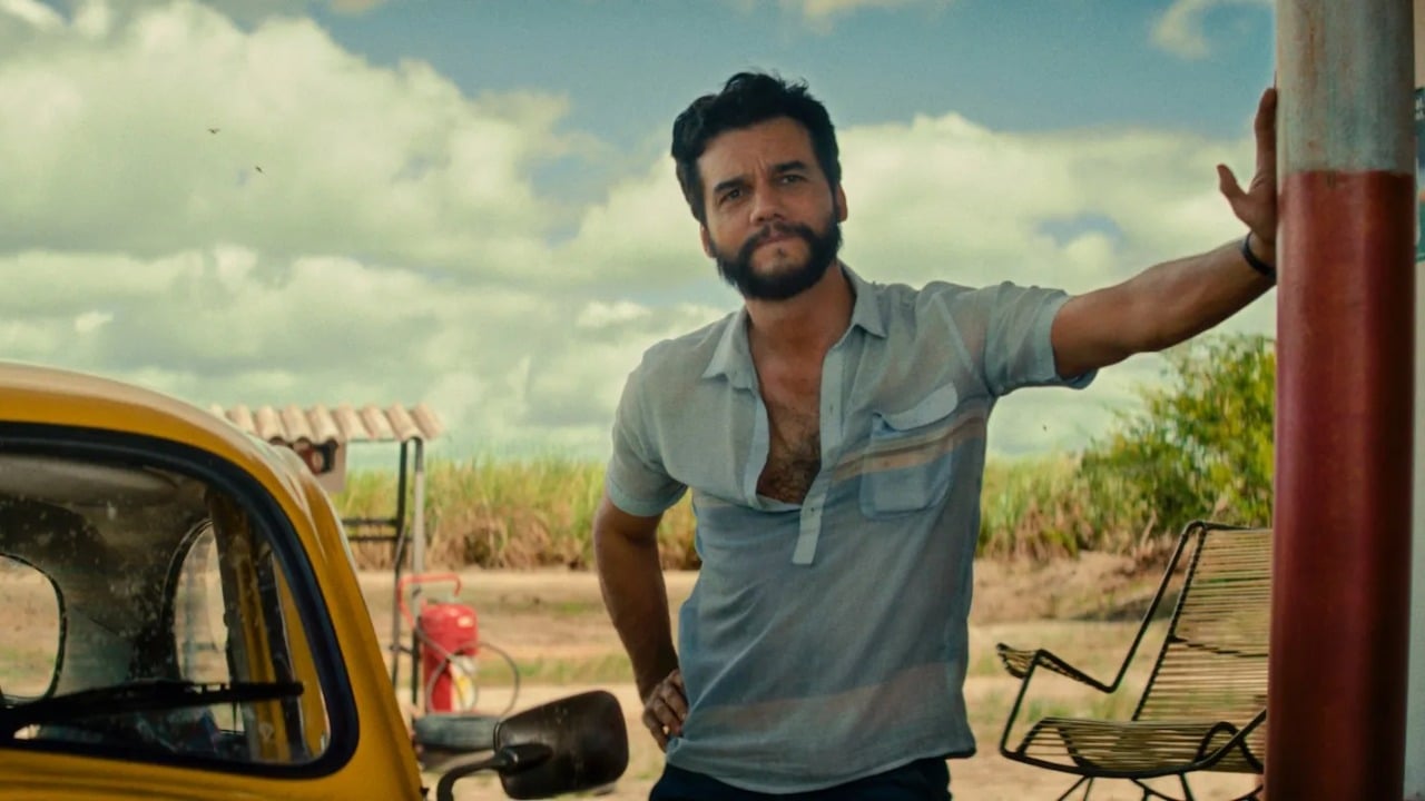 A man with a beard leans against a pole beside a yellow car, outdoors on a sunny day with grass, sky, and clouds in the background.