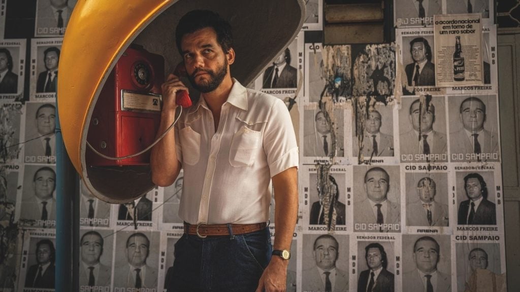 A man in a white shirt uses a public phone booth, standing in front of a wall covered with black-and-white posters of men's faces.