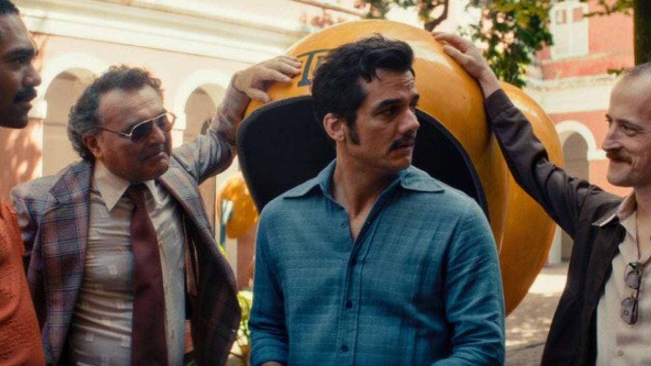 Four men stand together outside, with one man in a blue shirt in the center and a large yellow object behind them.