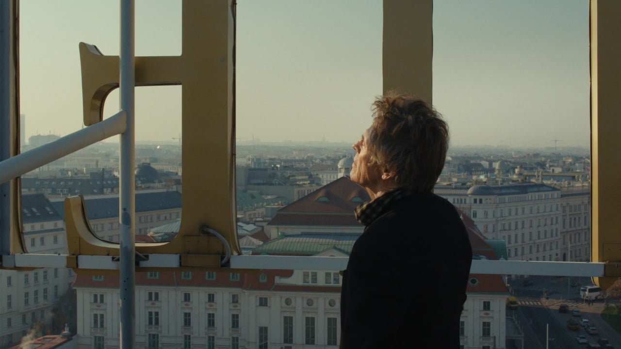 A man stands on a rooftop next to a large yellow letter "H," looking out over a cityscape with historic buildings under a clear sky.