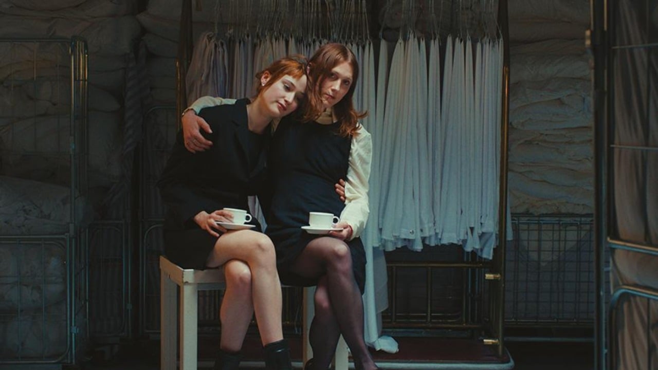 Two women sit closely together on a bench, each holding a teacup, with white linens and industrial carts in the background.