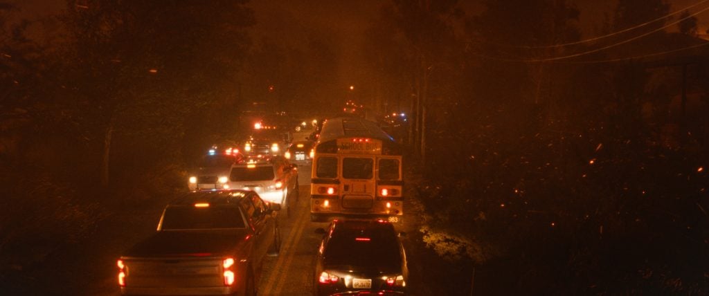 A line of cars and a school bus drive along a road at night, illuminated by headlights, with smoke or haze reducing visibility.
