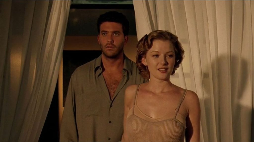 A man and woman stand indoors between white curtains; the man looks surprised, while the woman faces forward with a slight smile.