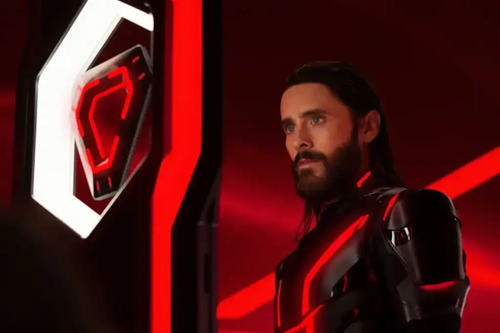 A man in a black, futuristic suit with red lights stands in a red-lit room, facing a white and red geometric display.