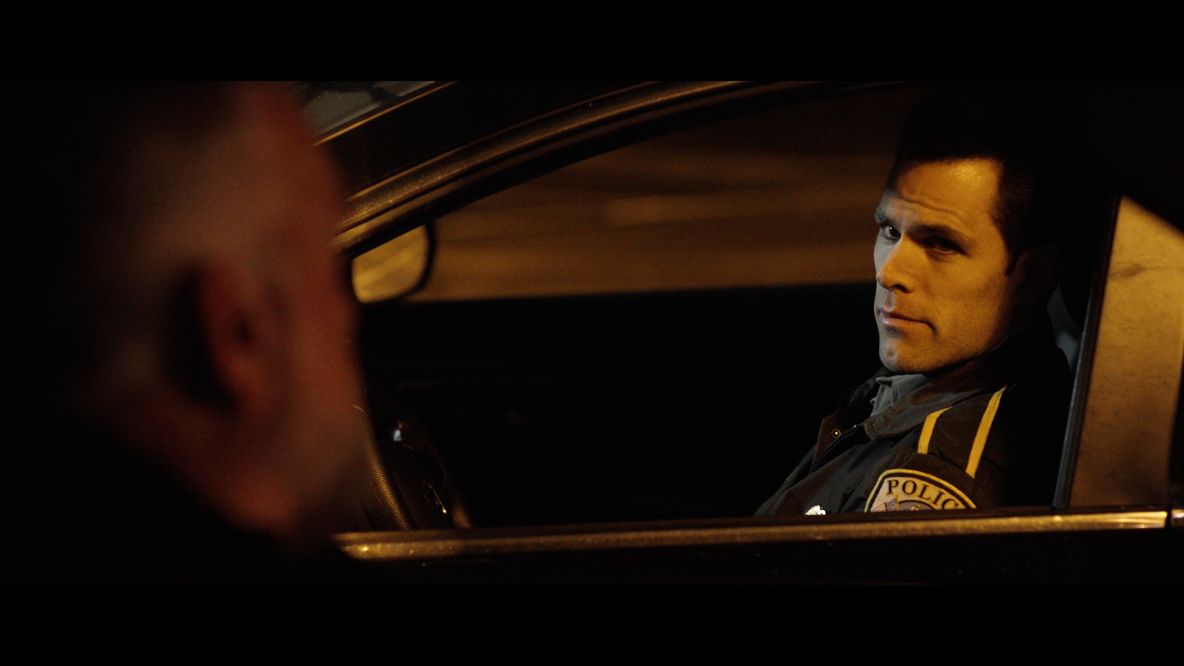 A police officer sits in a patrol car at night, looking out the window at a person who is partially visible in the foreground.