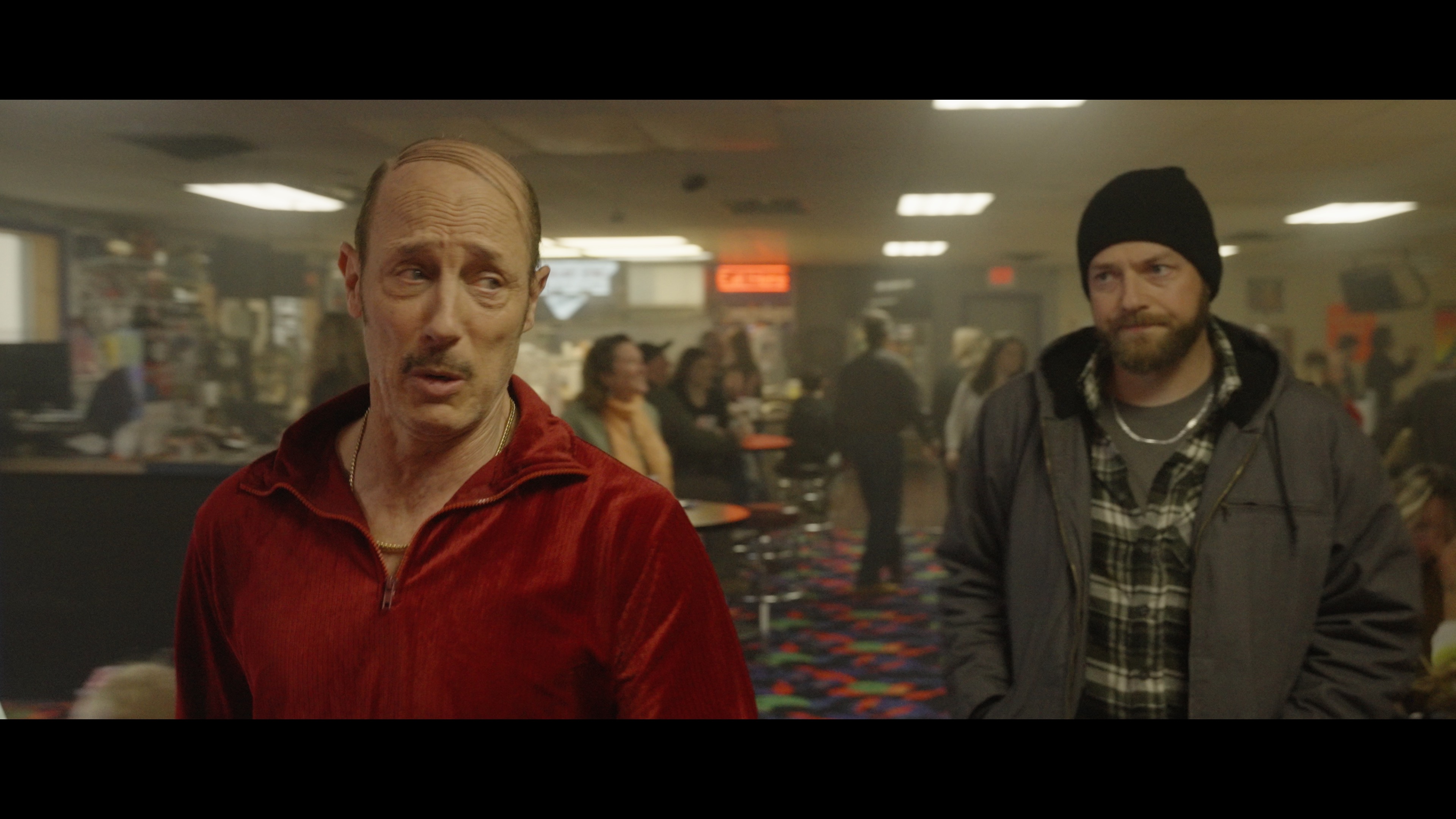 Two men stand in a bowling alley; the man in front wears a red velour tracksuit, while the man behind wears a beanie and plaid jacket. Other people are visible in the background.