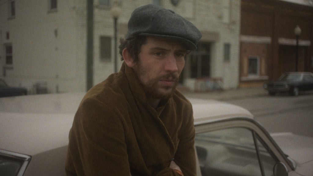 A man wearing a flat cap and brown jacket leans on the hood of a car parked on a street with buildings and a few parked cars in the background.