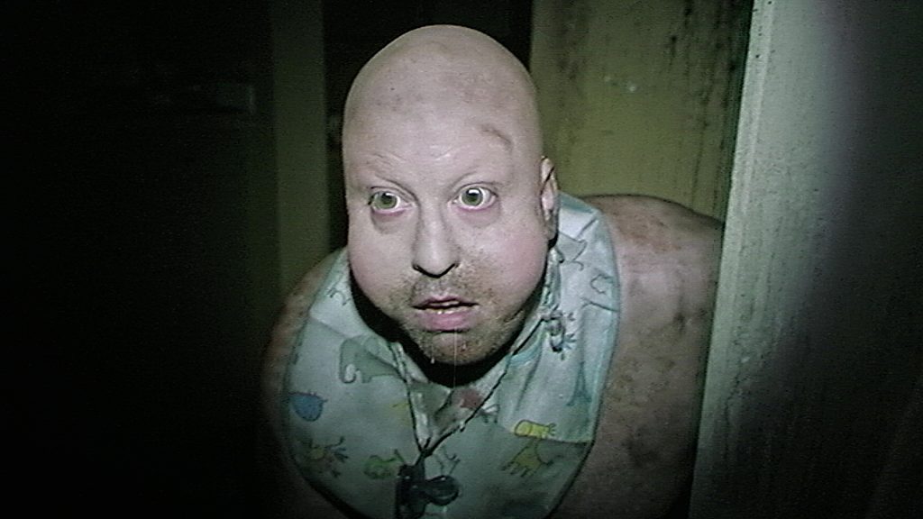 A bald person with pale skin, wide eyes, and a bib around their neck is drooling while peeking around a corner in a dimly lit setting.