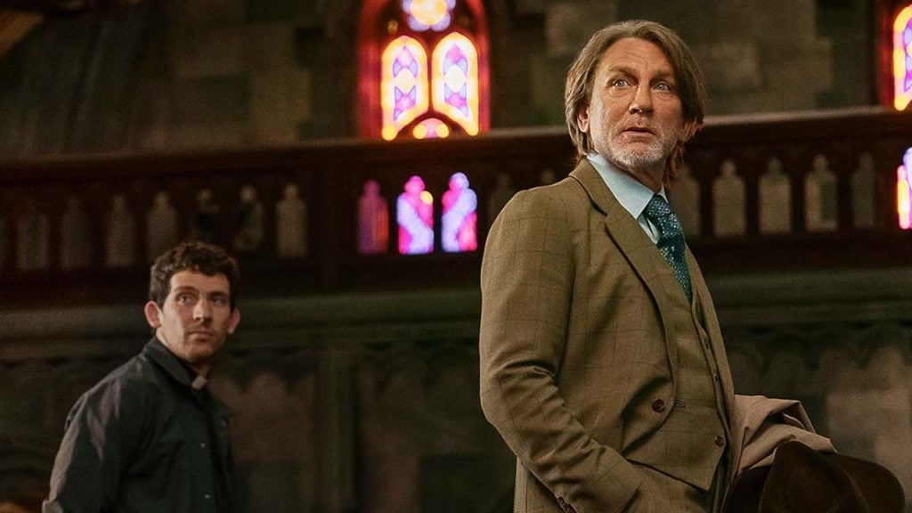 Two men stand inside a church with stained glass windows; one wears a brown suit and holds a coat, the other is dressed in dark clothing.