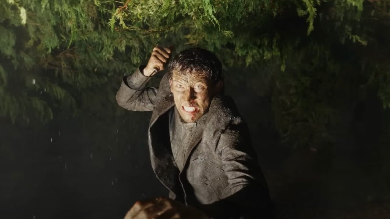 A man in a muddy suit holds a knife above his head, appearing intense and aggressive, with trees and darkness in the background.