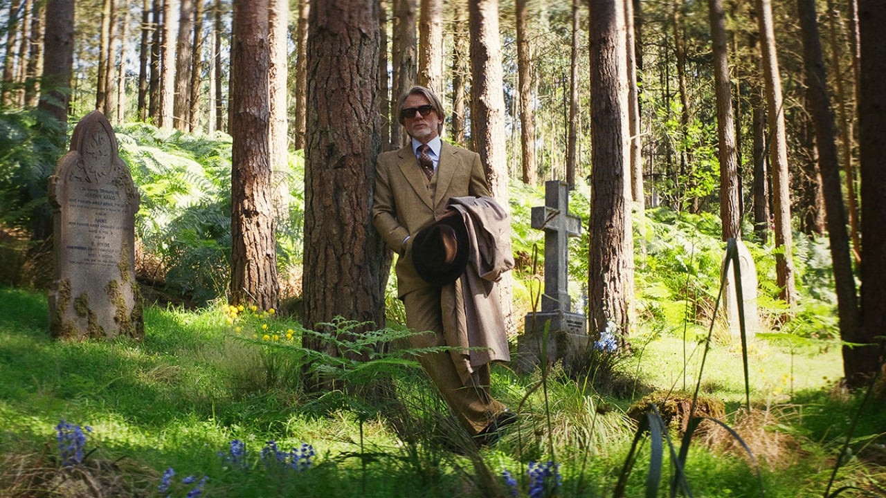An older man in a tan suit holding a coat walks through a sunlit forested cemetery with gravestones and lush greenery.