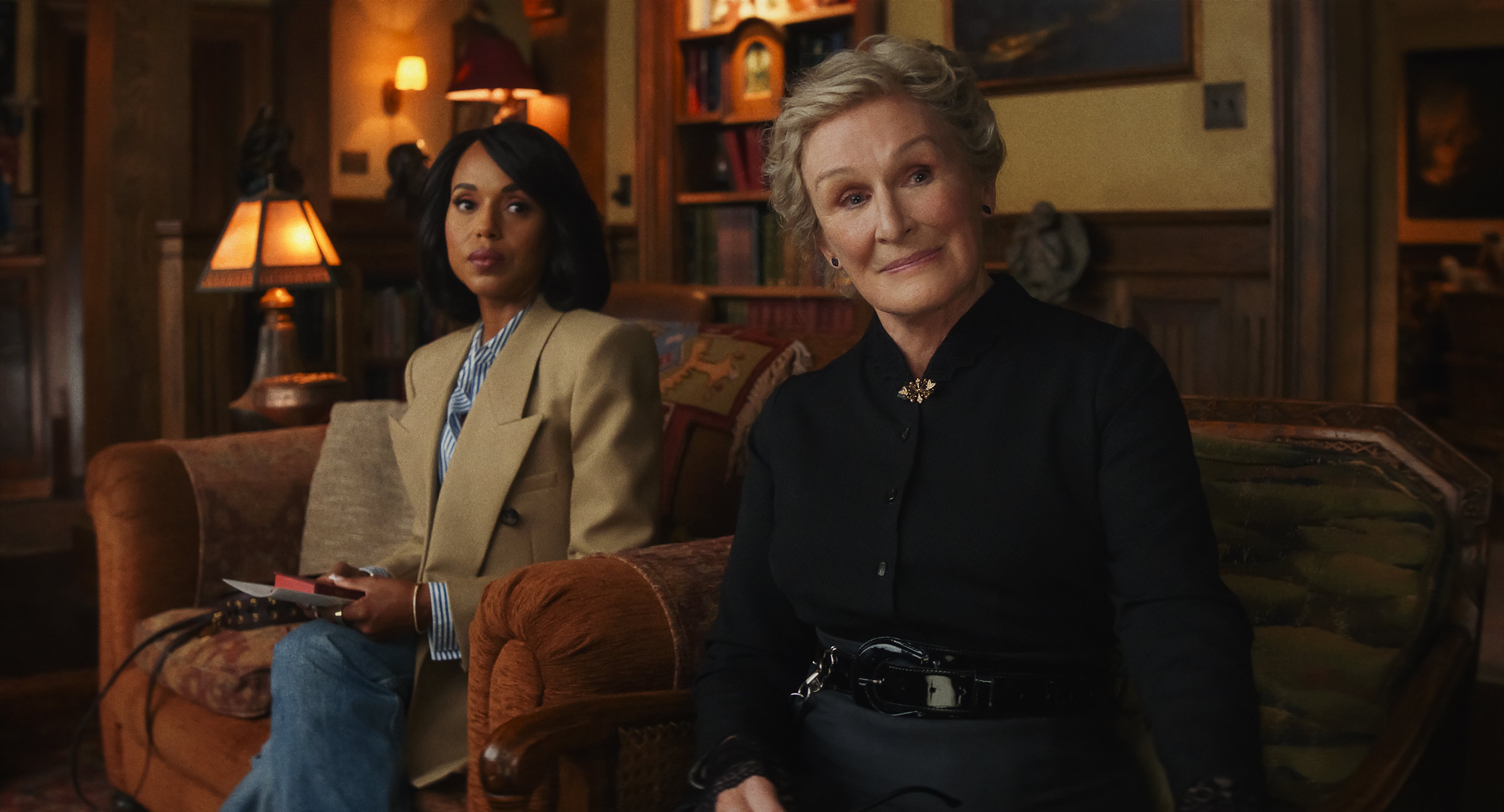 Two women sit in a warmly lit living room; one wears a tan blazer and holds papers, the other wears a black outfit and sits on a patterned sofa, both facing toward the camera.