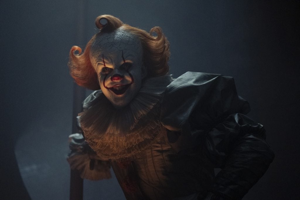 A person dressed as a sinister clown with white face makeup, red hair, and a ruffled costume stands in a dimly lit environment, holding a long object.