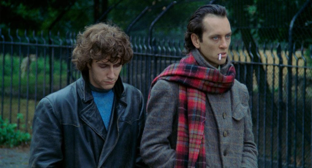 Two men walk outside near a black iron fence; one wears a leather jacket and looks down, the other wears a plaid scarf and coat with a cigarette in his mouth.
