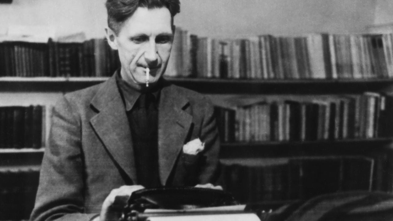A man in a suit sits at a typewriter with a cigarette in his mouth, surrounded by shelves of books in the background.