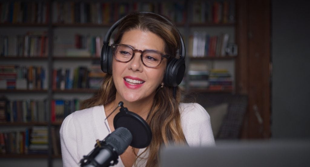 A woman wearing headphones and glasses speaks into a microphone, sitting in front of bookshelves.