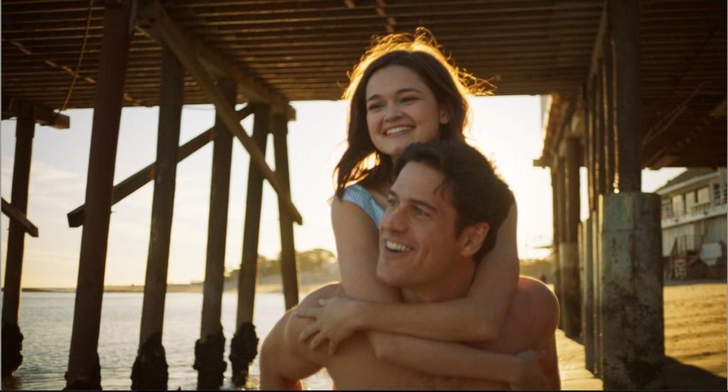 A woman smiles while riding on a man's back under a wooden pier at the beach during sunset.