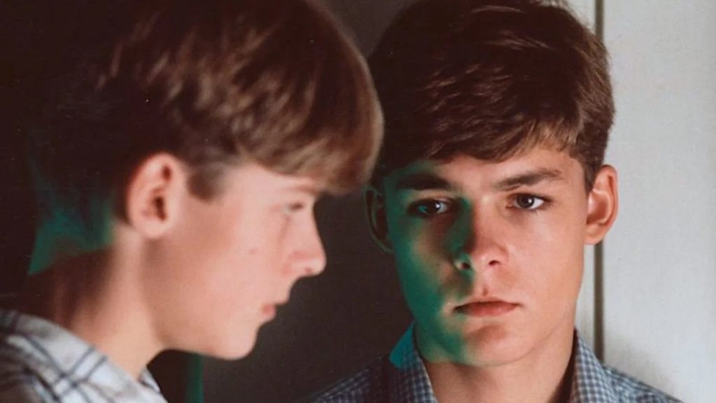 Two teenage boys stand close to each other indoors; one is facing forward with a serious expression while the other is shown in profile, slightly blurred.