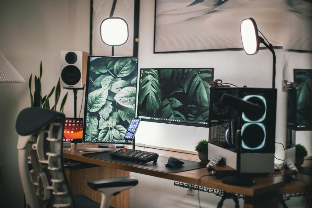 Modern computer desk setup with dual monitors displaying leaf images, a smartphone, ring lights, speakers, and a gaming PC in a well-lit room.
