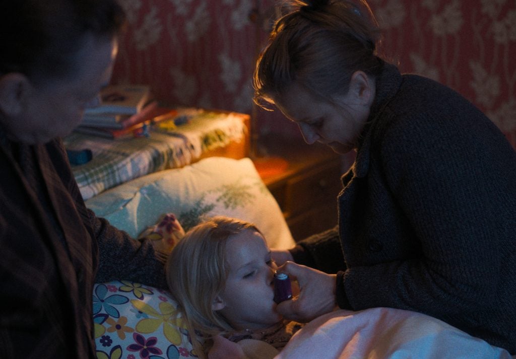 A woman helps a young girl in bed use an inhaler while another adult looks on in a softly lit bedroom.