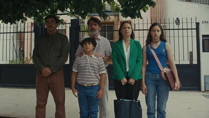 Five people stand in a line under a tree in front of a gated building, facing forward with neutral expressions.