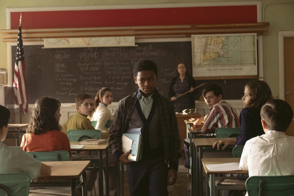 A student carrying books walks through a classroom as other students at their desks and a teacher at the chalkboard look toward him.