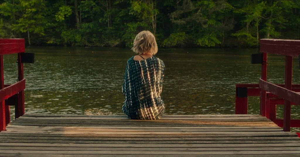 A person with short blonde hair sits alone on a wooden dock facing a calm lake, surrounded by green trees.