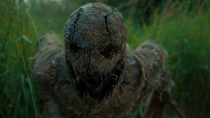 A figure dressed as a tattered, burlap-masked scarecrow stands in tall grass, with visible stitching and a gaping mouth hole on the mask.