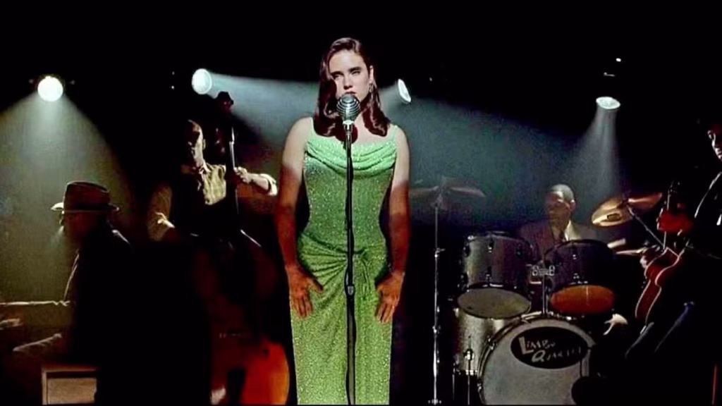 A woman in a green dress sings into a vintage microphone on stage, accompanied by a jazz band with a double bass, drum set, and guitar under stage lights.