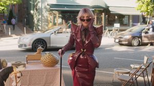 Universal Debuts Clip For Family Film ‘Gabby’s Dollhouse: The Movie’ To Celebrate Digital Release (EXCLUSIVE) A person in a shiny burgundy outfit with exaggerated shoulders and sunglasses stands on a sidewalk next to a table with baskets and a convertible car in the background.