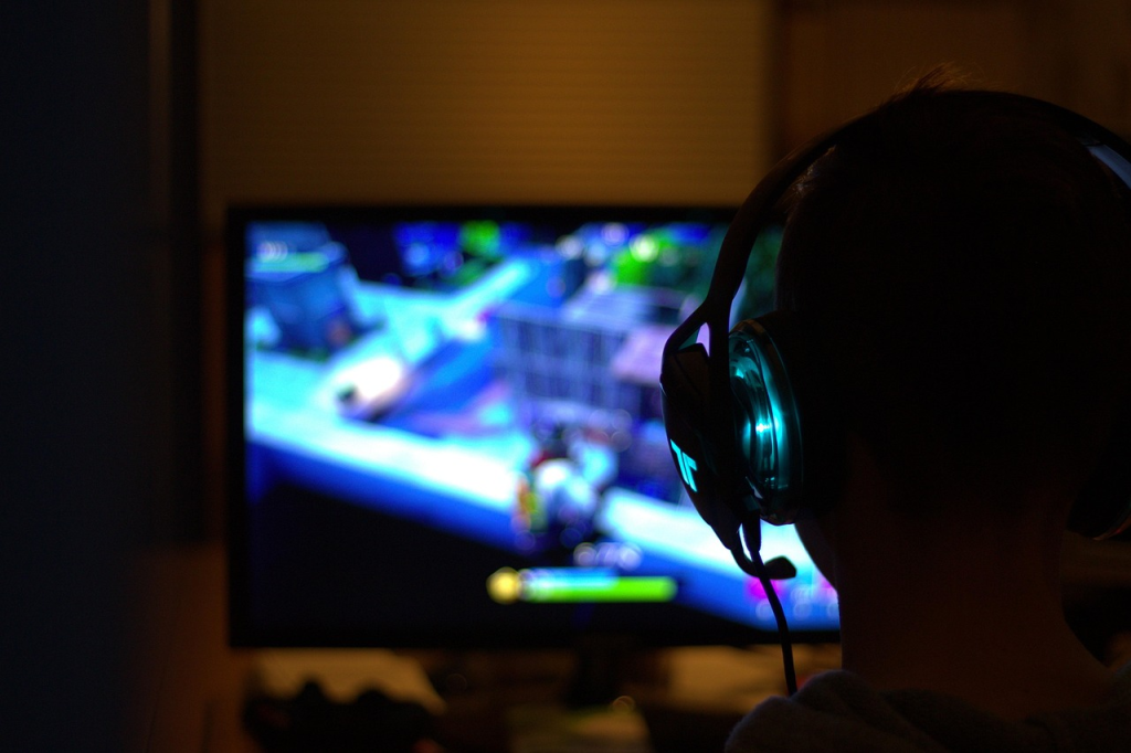Person wearing headphones sits in front of a monitor playing a colorful video game, with the screen showing game graphics in the background.