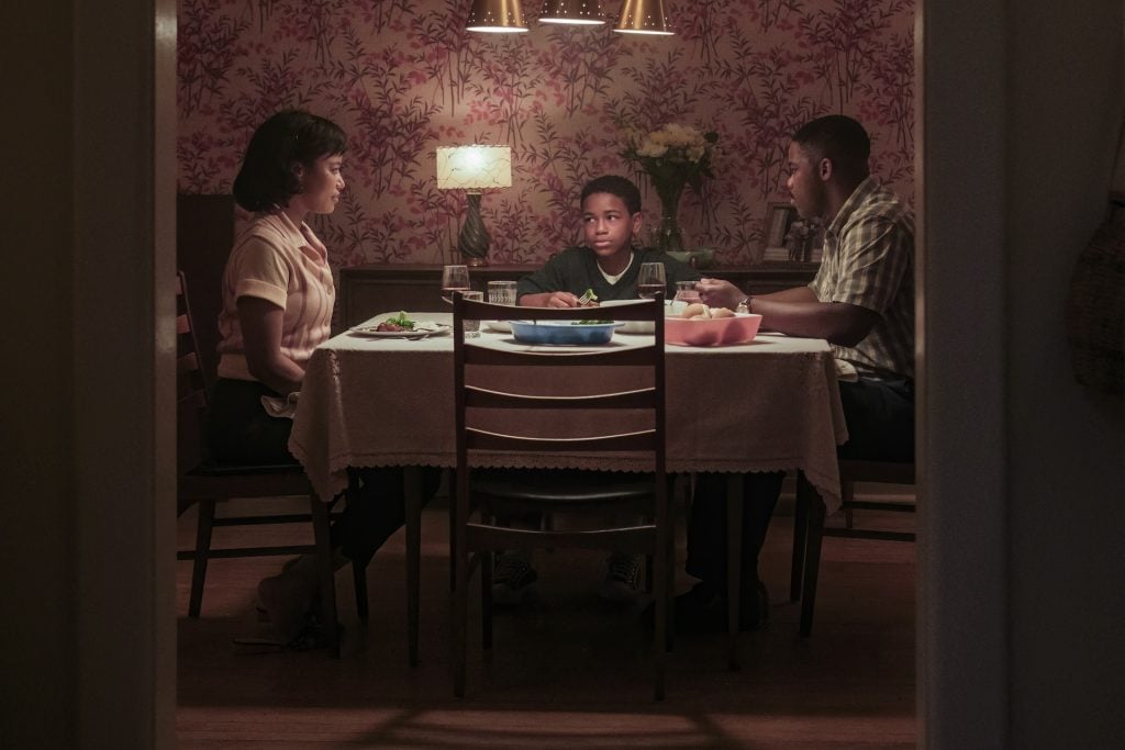 Three people sit silently around a dinner table in a dimly lit room with pink floral wallpaper, each with a plate of food in front of them.