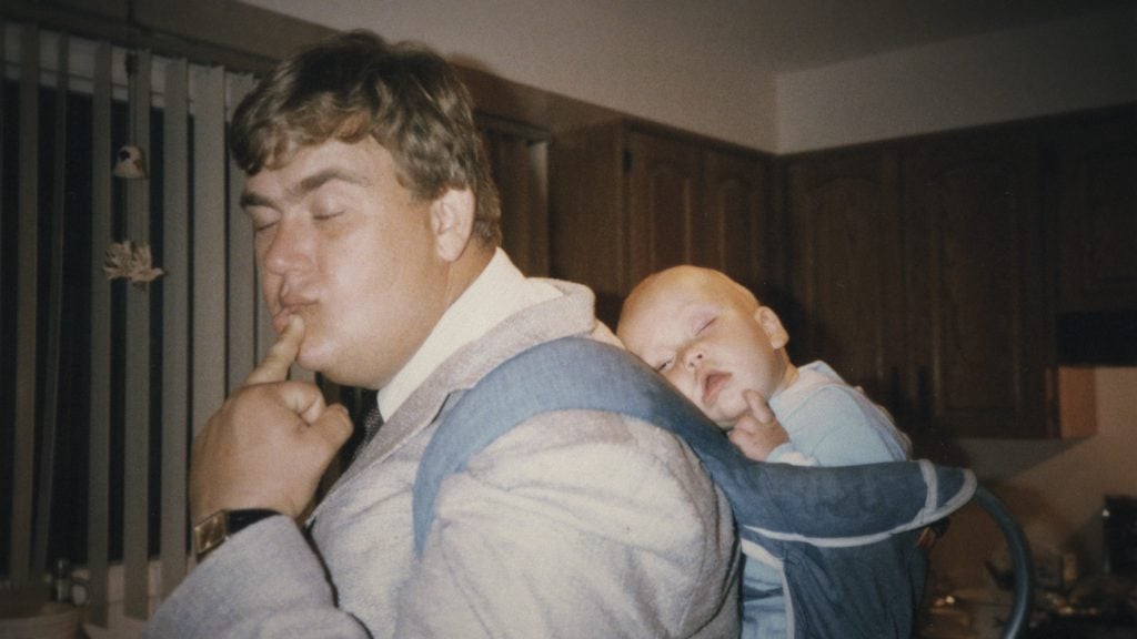 A man in a suit stands indoors, making a funny face with his finger on his lips, while a sleeping baby rests in a blue carrier on his back.