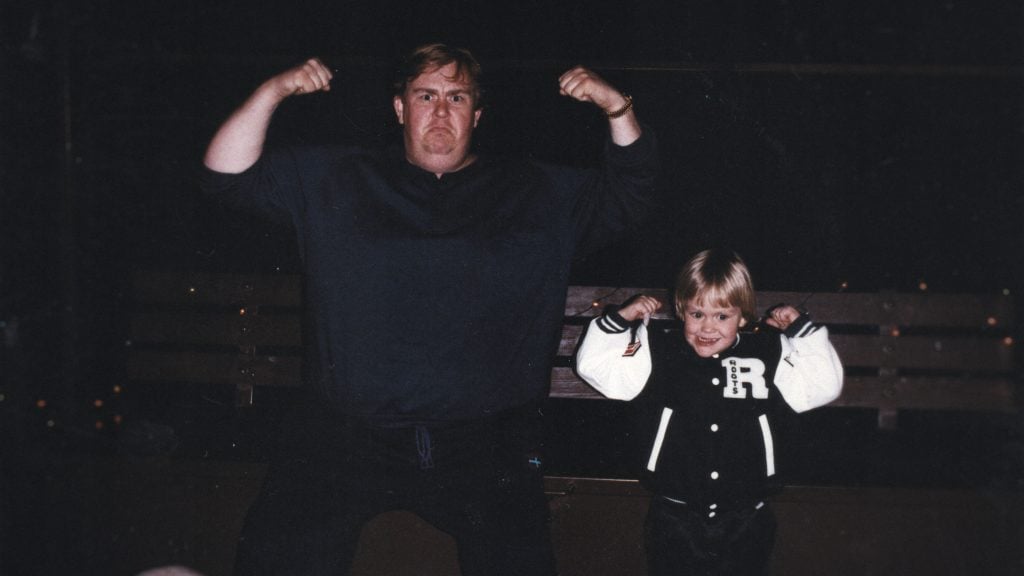 An adult and a child pose side by side, both flexing their arms and making strong expressions; the child wears a black and white letterman jacket.